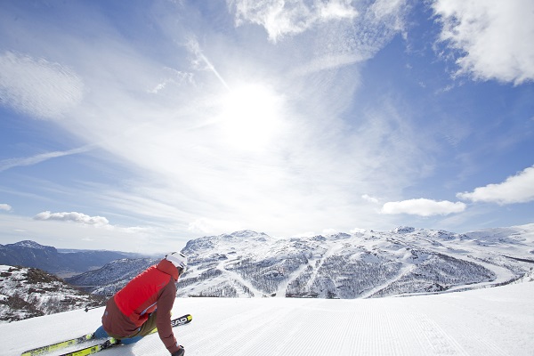 Person som ker skidor i en skidbacke. Sntckta backar, bl himmel och sol.